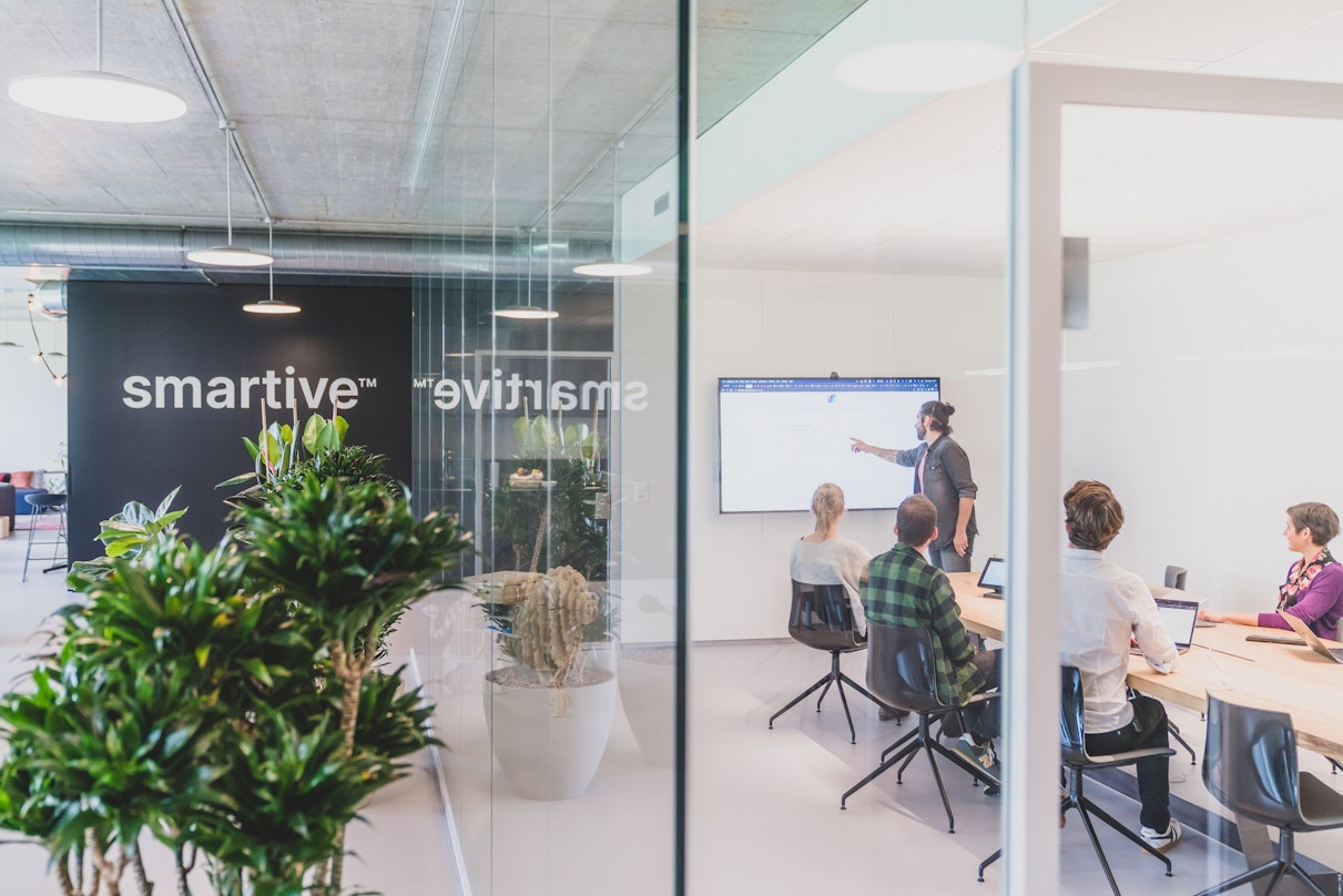 Ein Mann steht und präsentiert drei sitzenden Kollegen in einem modernen Büro mit Glaswänden und Grünpflanzen Informationen auf einem Bildschirm. An der Wand hinter ihnen ist das Wort „smartive“ zu sehen.