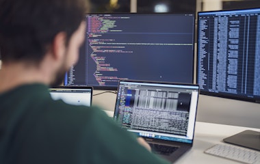 Eine Person arbeitet an einem Schreibtisch mit mehreren Monitoren, auf denen Code und Daten angezeigt werden.