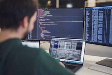 Eine Person arbeitet an einem Schreibtisch mit mehreren Monitoren, auf denen Code und Daten angezeigt werden.