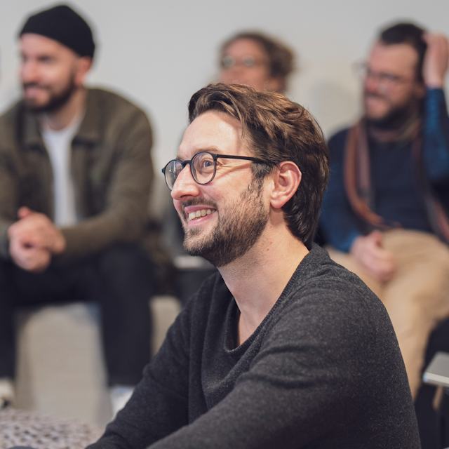 Ein Mann sitzt lächelnd auf dem Boden in einem Raum mit mehreren anderen Personen, die auf einem Sofa im Hintergrund sitzen und ebenfalls lachen.