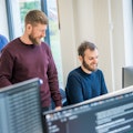 Zwei Männer unterhalten sich und lächeln vor einem Computerbildschirm in einem Büro, während im Hintergrund eine weitere Person arbeitet.