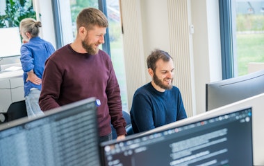 Zwei Männer unterhalten sich und lächeln vor einem Computerbildschirm in einem Büro, während im Hintergrund eine weitere Person arbeitet.