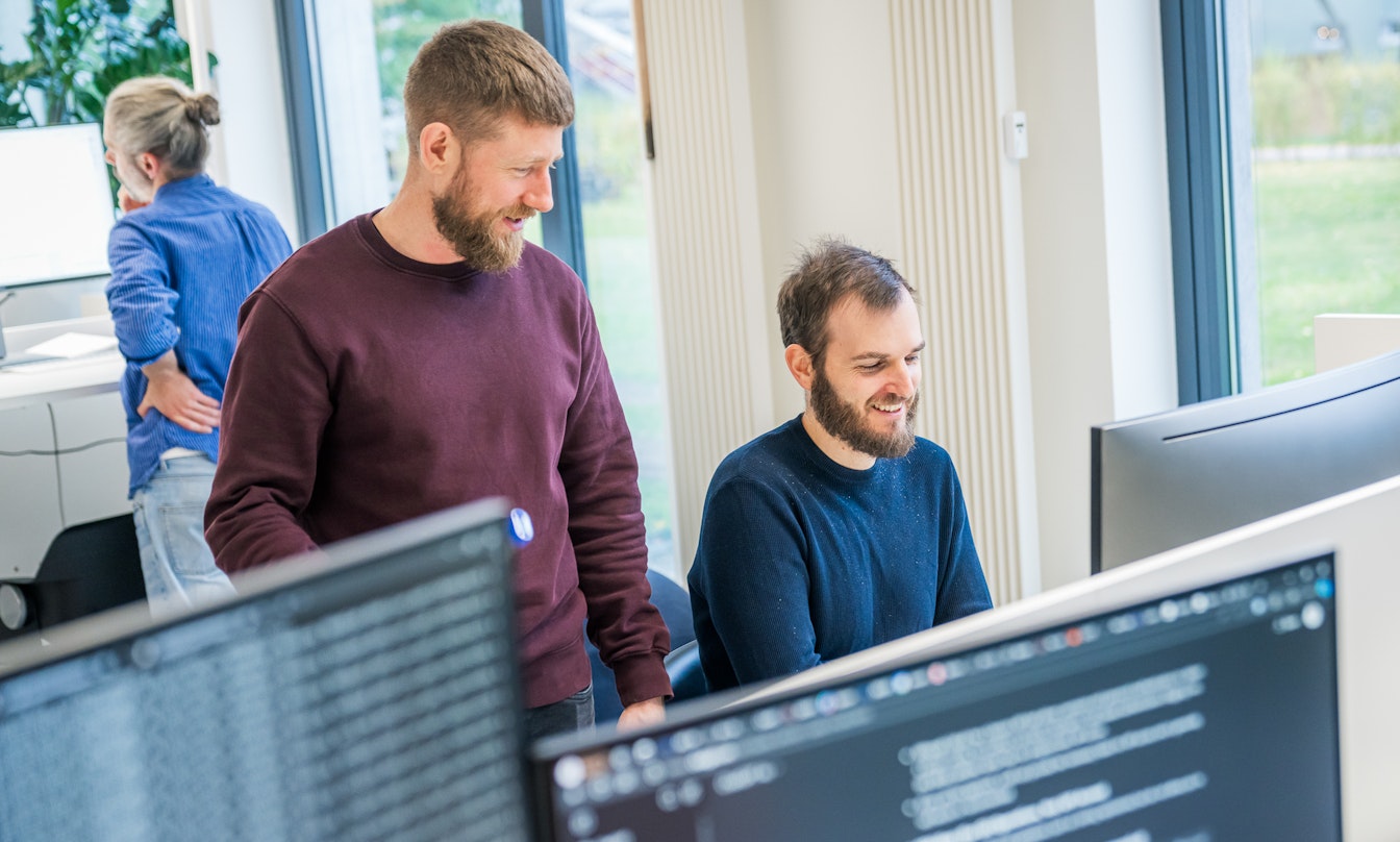 Zwei Männer unterhalten sich und lächeln vor einem Computerbildschirm in einem Büro, während im Hintergrund eine weitere Person arbeitet.