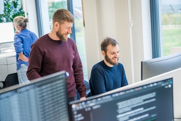 Zwei Männer unterhalten sich und lächeln vor einem Computerbildschirm in einem Büro, während im Hintergrund eine weitere Person arbeitet.
