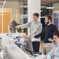 Zwei Männer stehen an einem Schreibtisch und schauen auf einen Computerbildschirm in einem Großraumbüro. Eine weitere Person sitzt mit Kopfhörern an einem Schreibtisch im Vordergrund.