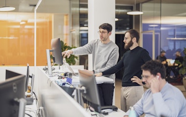 Zwei Männer stehen an einem Schreibtisch und schauen auf einen Computerbildschirm in einem Großraumbüro. Eine weitere Person sitzt mit Kopfhörern an einem Schreibtisch im Vordergrund.