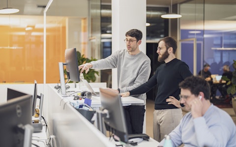 Zwei Männer stehen an einem Schreibtisch und schauen auf einen Computerbildschirm in einem Großraumbüro. Eine weitere Person sitzt mit Kopfhörern an einem Schreibtisch im Vordergrund.