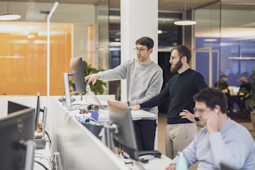 Zwei Männer stehen an einem Schreibtisch und schauen auf einen Computerbildschirm in einem Großraumbüro. Eine weitere Person sitzt mit Kopfhörern an einem Schreibtisch im Vordergrund.