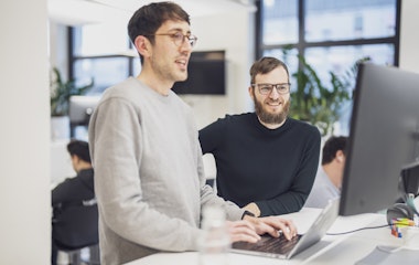 Zwei Mitarbeitende stehen an einem Schreibtisch im Büro. Eine tippt auf einem Laptop, während die andere auf den Bildschirm schaut.