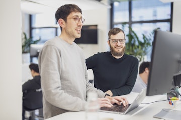 Zwei Mitarbeitende stehen an einem Schreibtisch im Büro. Eine tippt auf einem Laptop, während die andere auf den Bildschirm schaut.