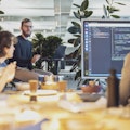 Eine Gruppe von Menschen in einem modernen Büro hält eine Präsentation. Auf einem großen Bildschirm wird Computercode angezeigt. Pflanzen und Büromaterial sind zu sehen.