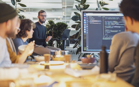 Eine Gruppe von Menschen in einem modernen Büro hält eine Präsentation. Auf einem großen Bildschirm wird Computercode angezeigt. Pflanzen und Büromaterial sind zu sehen.
