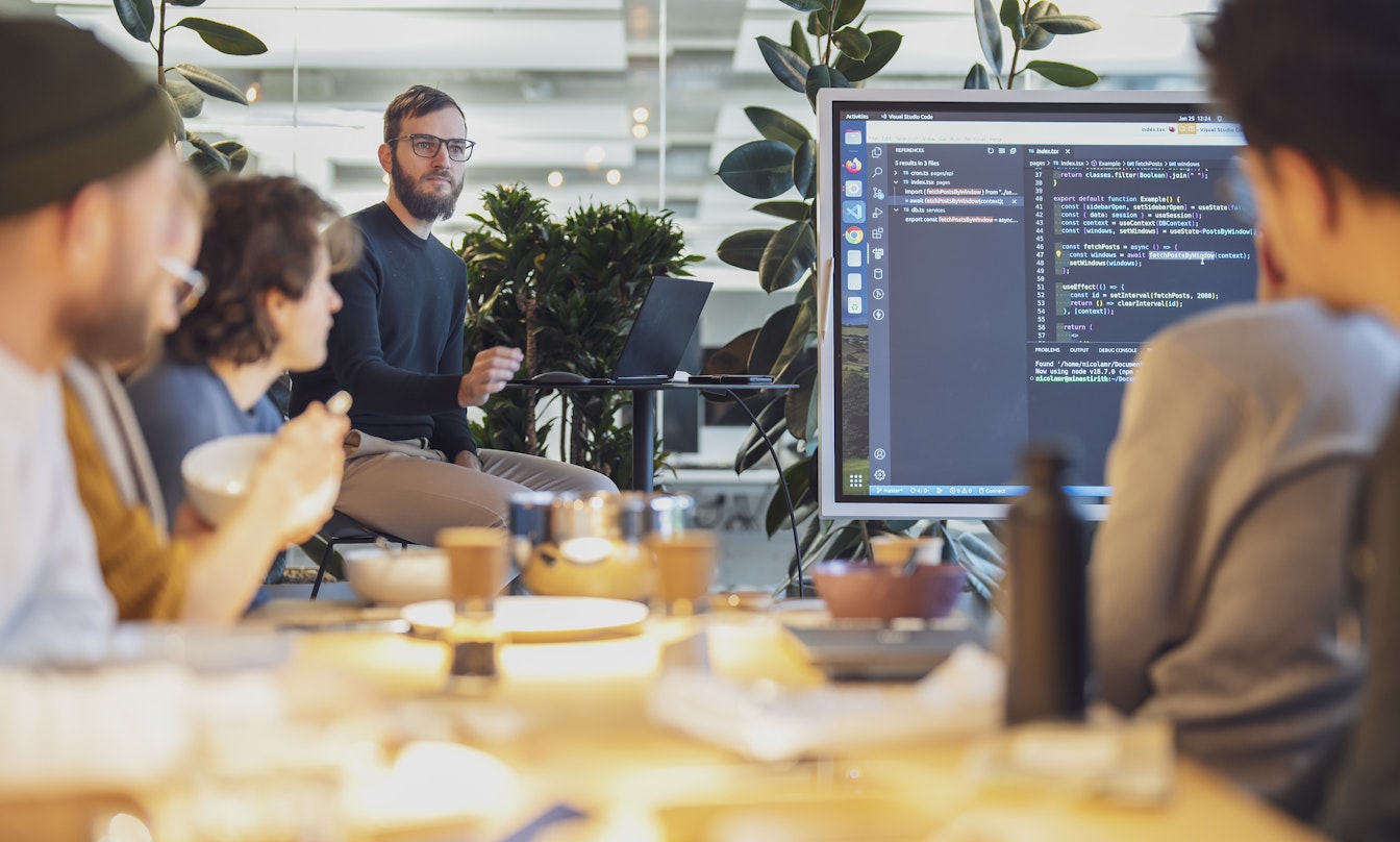 Eine Gruppe von Menschen in einem modernen Büro hält eine Präsentation. Auf einem großen Bildschirm wird Computercode angezeigt. Pflanzen und Büromaterial sind zu sehen.