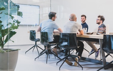 Vier Personen sitzen in einem Konferenzraum mit Glaswänden um einen Konferenztisch herum. Im Hintergrund zeigt ein grosser Bildschirm Informationen an.