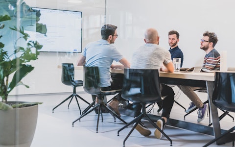 Vier Personen sitzen in einem Konferenzraum mit Glaswänden um einen Konferenztisch herum. Im Hintergrund zeigt ein grosser Bildschirm Informationen an.