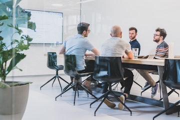 Vier Personen sitzen in einem Konferenzraum mit Glaswänden um einen Konferenztisch herum. Im Hintergrund zeigt ein grosser Bildschirm Informationen an.