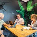 Drei Personen unterhalten sich an einem Holztisch in einem Raum mit grünen Blattgemälden und einer Affenskulptur. Zwei haben Laptops geöffnet, während der dritte eine Tasse hält.