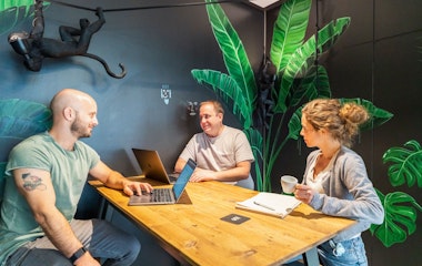 Drei Personen unterhalten sich an einem Holztisch in einem Raum mit grünen Blattgemälden und einer Affenskulptur. Zwei haben Laptops geöffnet, während der dritte eine Tasse hält.