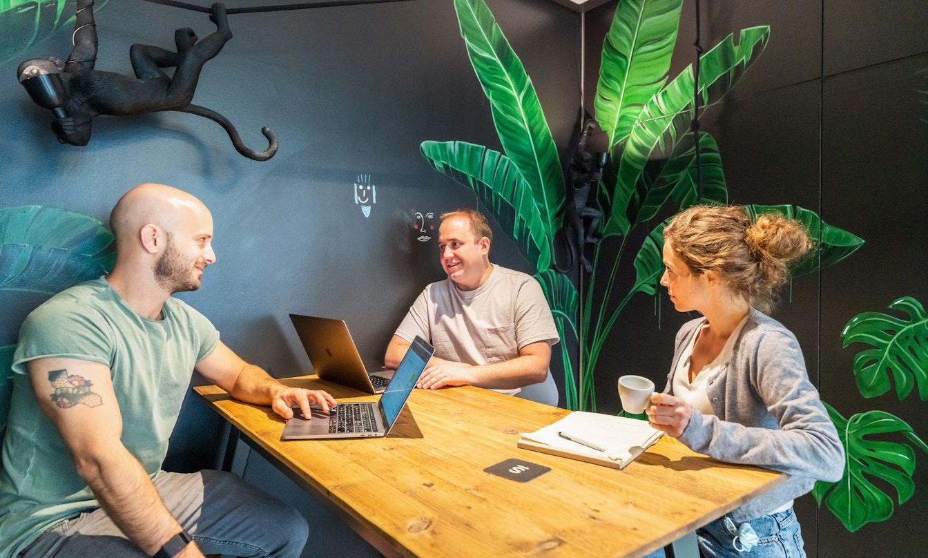 Drei Personen unterhalten sich an einem Holztisch in einem Raum mit grünen Blattgemälden und einer Affenskulptur. Zwei haben Laptops geöffnet, während der dritte eine Tasse hält.