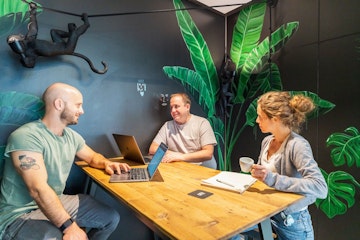 Drei Personen unterhalten sich an einem Holztisch in einem Raum mit grünen Blattgemälden und einer Affenskulptur. Zwei haben Laptops geöffnet, während der dritte eine Tasse hält.