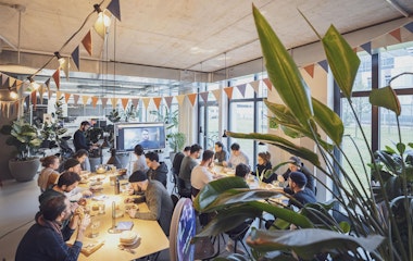 Eine Gruppe von Menschen sitzt in einem modernen, gut beleuchteten Raum an Tischen und sieht sich eine Präsentation auf einer Leinwand an. Der Raum ist mit dreieckigen Fahnen und Pflanzen dekoriert.