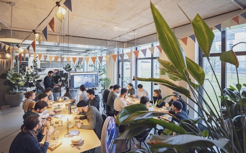 Eine Gruppe von Menschen sitzt in einem modernen, gut beleuchteten Raum an Tischen und sieht sich eine Präsentation auf einer Leinwand an. Der Raum ist mit dreieckigen Fahnen und Pflanzen dekoriert.