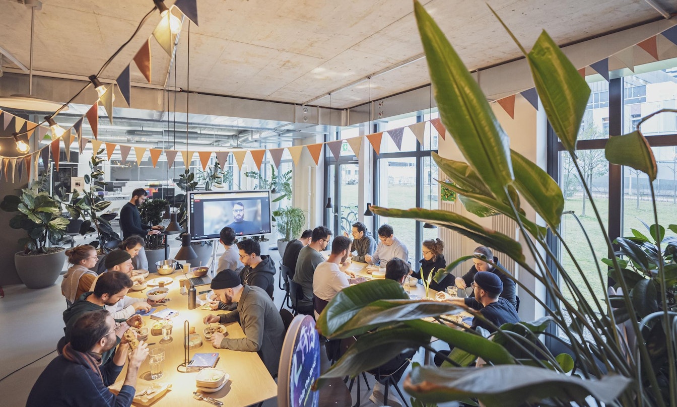 Eine Gruppe von Menschen sitzt in einem modernen, gut beleuchteten Raum an Tischen und sieht sich eine Präsentation auf einer Leinwand an. Der Raum ist mit dreieckigen Fahnen und Pflanzen dekoriert.