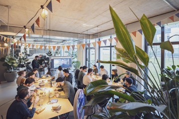 Eine Gruppe von Menschen sitzt in einem modernen, gut beleuchteten Raum an Tischen und sieht sich eine Präsentation auf einer Leinwand an. Der Raum ist mit dreieckigen Fahnen und Pflanzen dekoriert.