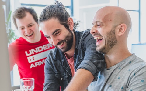 Drei Männer lächeln und lachen zusammen in einem Gebäude, wobei einer dem anderen über die Schulter greift.