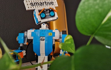 Lego-Roboter mit 'smartive'-Schild im Hintergrund, umgeben von grünen Pflanzen.