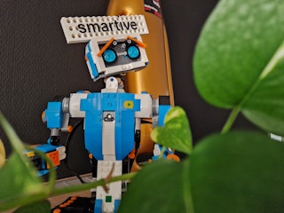 Lego-Roboter mit 'smartive'-Schild im Hintergrund, umgeben von grünen Pflanzen.