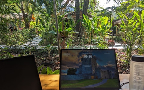 Laptops und eine Wasserflasche auf einem Holztisch mit einem üppigen Garten im Hintergrund.