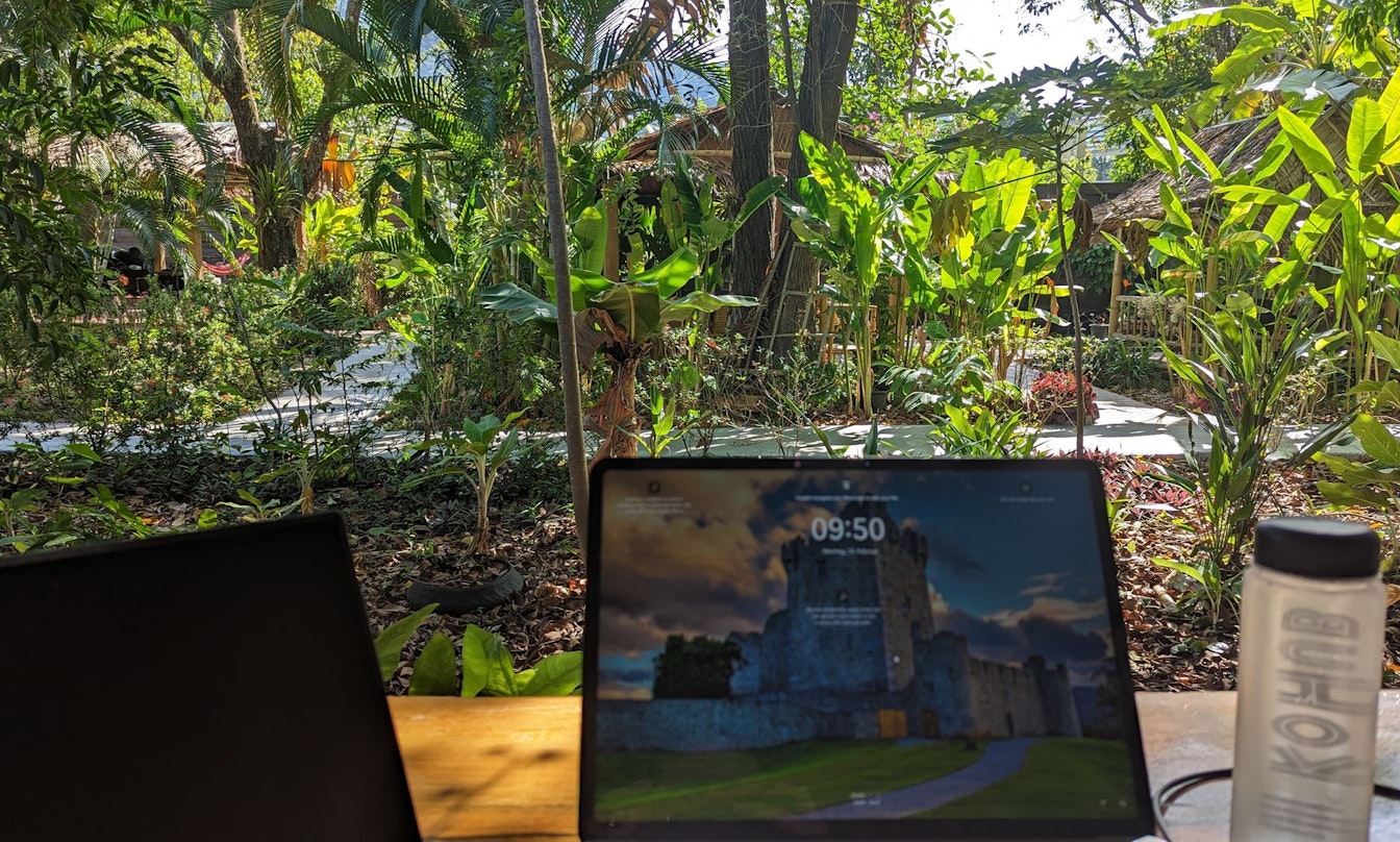 Laptops und eine Wasserflasche auf einem Holztisch mit einem üppigen Garten im Hintergrund.