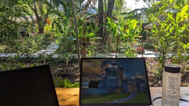 Laptops und eine Wasserflasche auf einem Holztisch mit einem üppigen Garten im Hintergrund.