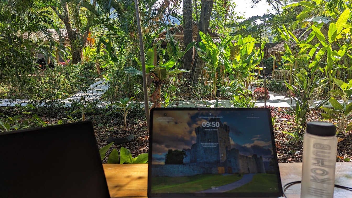 Laptops und eine Wasserflasche auf einem Holztisch mit einem üppigen Garten im Hintergrund.