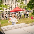 Menschen spielen im Freien in einer parkähnlichen Umgebung Tischtennis, mit einer Menschenmenge und einem roten Baldachin-Zelt im Hintergrund.