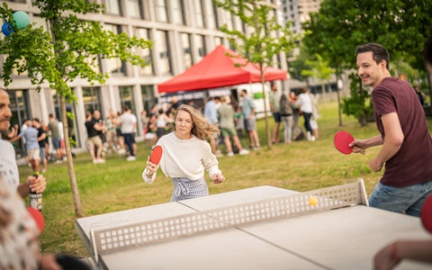 Menschen spielen im Freien in einer parkähnlichen Umgebung Tischtennis, mit einer Menschenmenge und einem roten Baldachin-Zelt im Hintergrund.