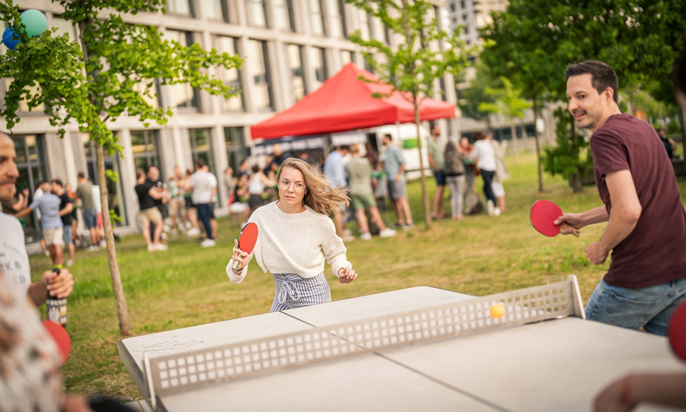 Menschen spielen im Freien in einer parkähnlichen Umgebung Tischtennis, mit einer Menschenmenge und einem roten Baldachin-Zelt im Hintergrund.