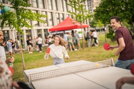 Menschen spielen im Freien in einer parkähnlichen Umgebung Tischtennis, mit einer Menschenmenge und einem roten Baldachin-Zelt im Hintergrund.