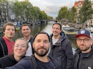 Eine Gruppe von sechs Personen macht ein Selfie auf einer Brücke mit einem Kanal und Gebäuden im Hintergrund.