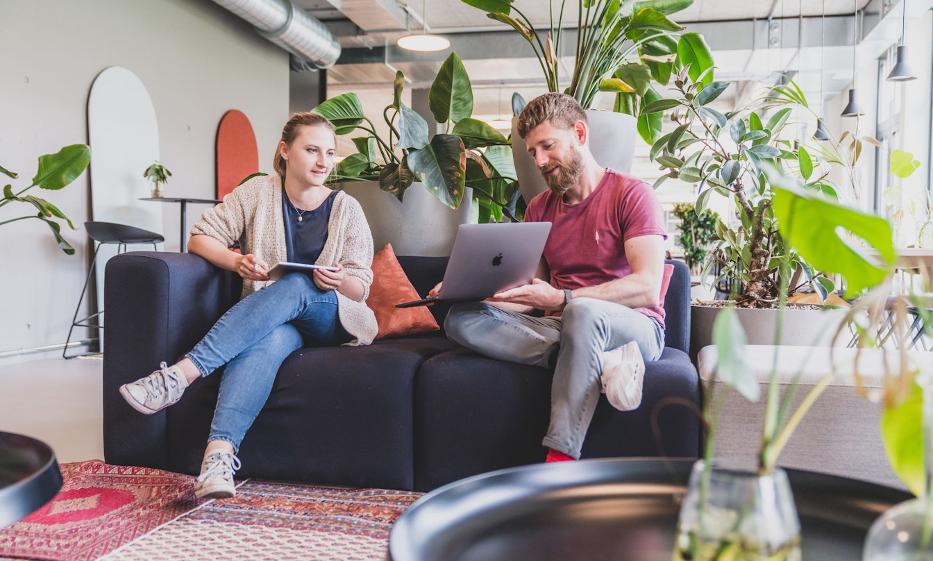 In einem modernen Büro mit Pflanzen sitzen zwei Personen auf einer Couch, beide mit einem Laptop und einem Tablet, und unterhalten sich.