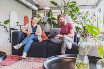 In einem modernen Büro mit Pflanzen sitzen zwei Personen auf einer Couch, beide mit einem Laptop und einem Tablet, und unterhalten sich.