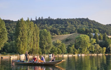 Eine Gruppe von Menschen in einem Holzboot auf einem ruhigen See, umgeben von grünen Bäumen und Hügeln unter einem klaren blauen Himmel.