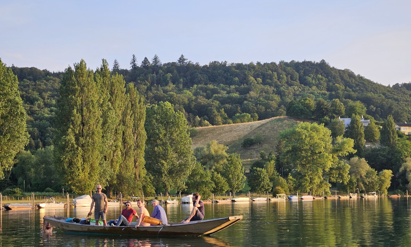 Eine Gruppe von Menschen in einem Holzboot auf einem ruhigen See, umgeben von grünen Bäumen und Hügeln unter einem klaren blauen Himmel.