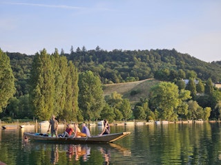 Eine Gruppe von Menschen in einem Holzboot auf einem ruhigen See, umgeben von grünen Bäumen und Hügeln unter einem klaren blauen Himmel.