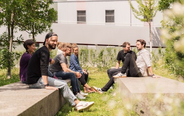 Eine Gruppe von Menschen sitzt auf Steinbänken in einem grasbewachsenen Außenbereich, einige einander gegenüber, mit Bäumen und einem Gebäude im Hintergrund.