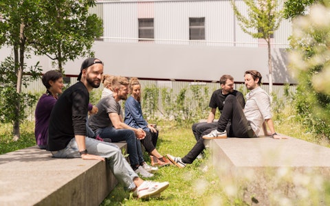 Eine Gruppe von Menschen sitzt auf Steinbänken in einem grasbewachsenen Außenbereich, einige einander gegenüber, mit Bäumen und einem Gebäude im Hintergrund.