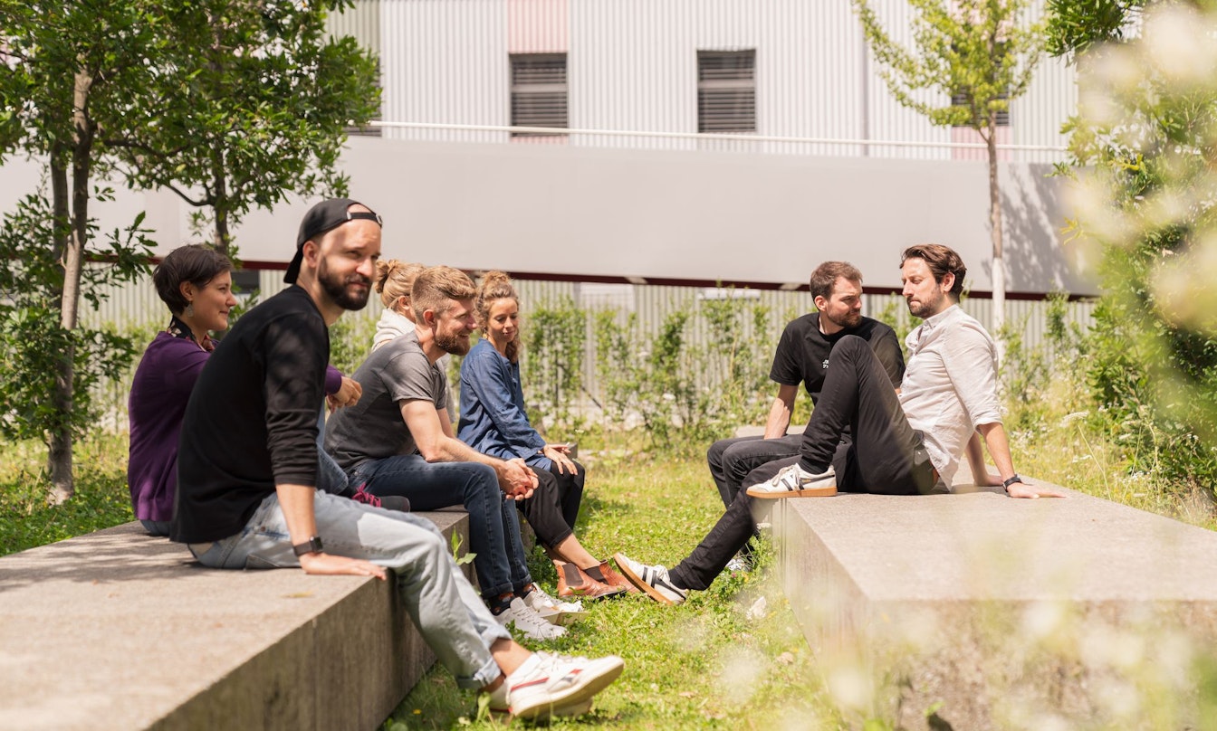 Eine Gruppe von Menschen sitzt auf Steinbänken in einem grasbewachsenen Außenbereich, einige einander gegenüber, mit Bäumen und einem Gebäude im Hintergrund.