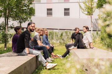 Eine Gruppe von Menschen sitzt auf Steinbänken in einem grasbewachsenen Außenbereich, einige einander gegenüber, mit Bäumen und einem Gebäude im Hintergrund.
