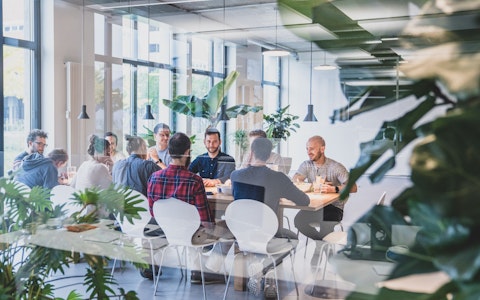 Eine Gruppe von Menschen sitzt um einen großen Tisch in einem modernen Büroraum mit Pflanzen und Deckenbeleuchtung.